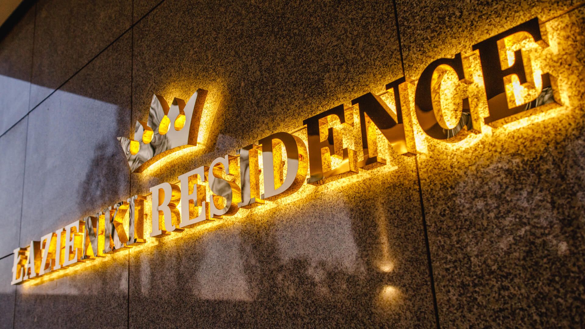 Łazienki Residence - Napis Łazienki Residence wykonany z blachy nierdzewnej w kolorze złotym, podświetlane LED tyłem na ścianę, z koroną w logo.