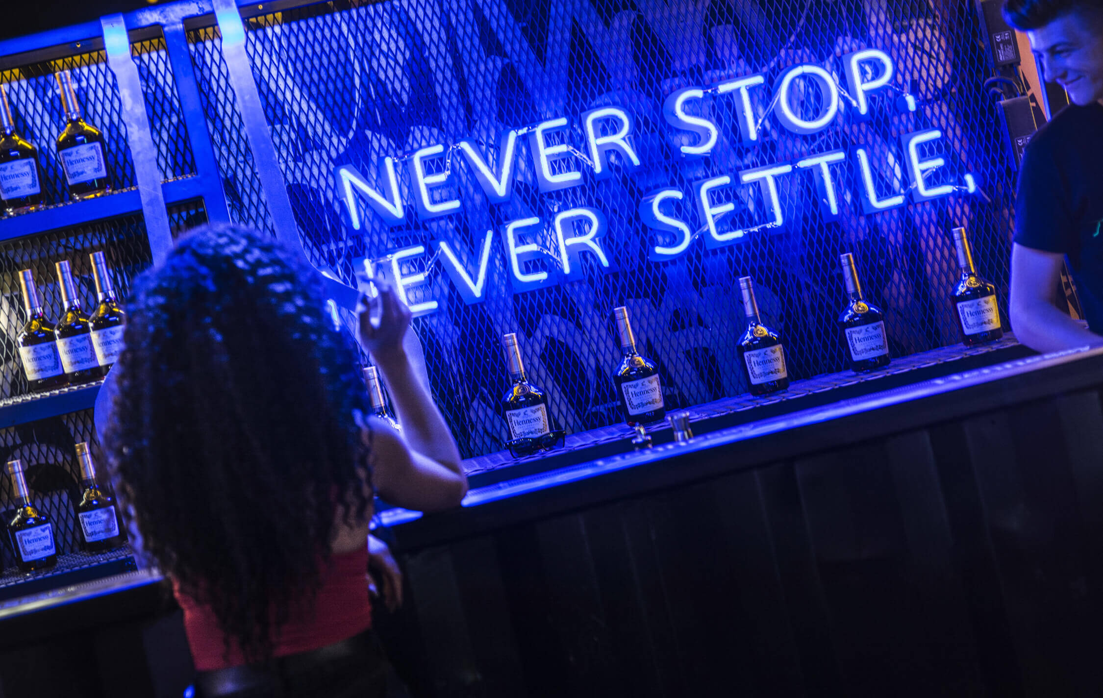 nie-aufhören-zu-siedeln-Hennessy-Neon-Werbung-Hennessy-Neon-Schriftzug-Neon-Aufschrift-auf-der-Brust-der-Flaschen-Blau-Neon-hinter-der-Bar