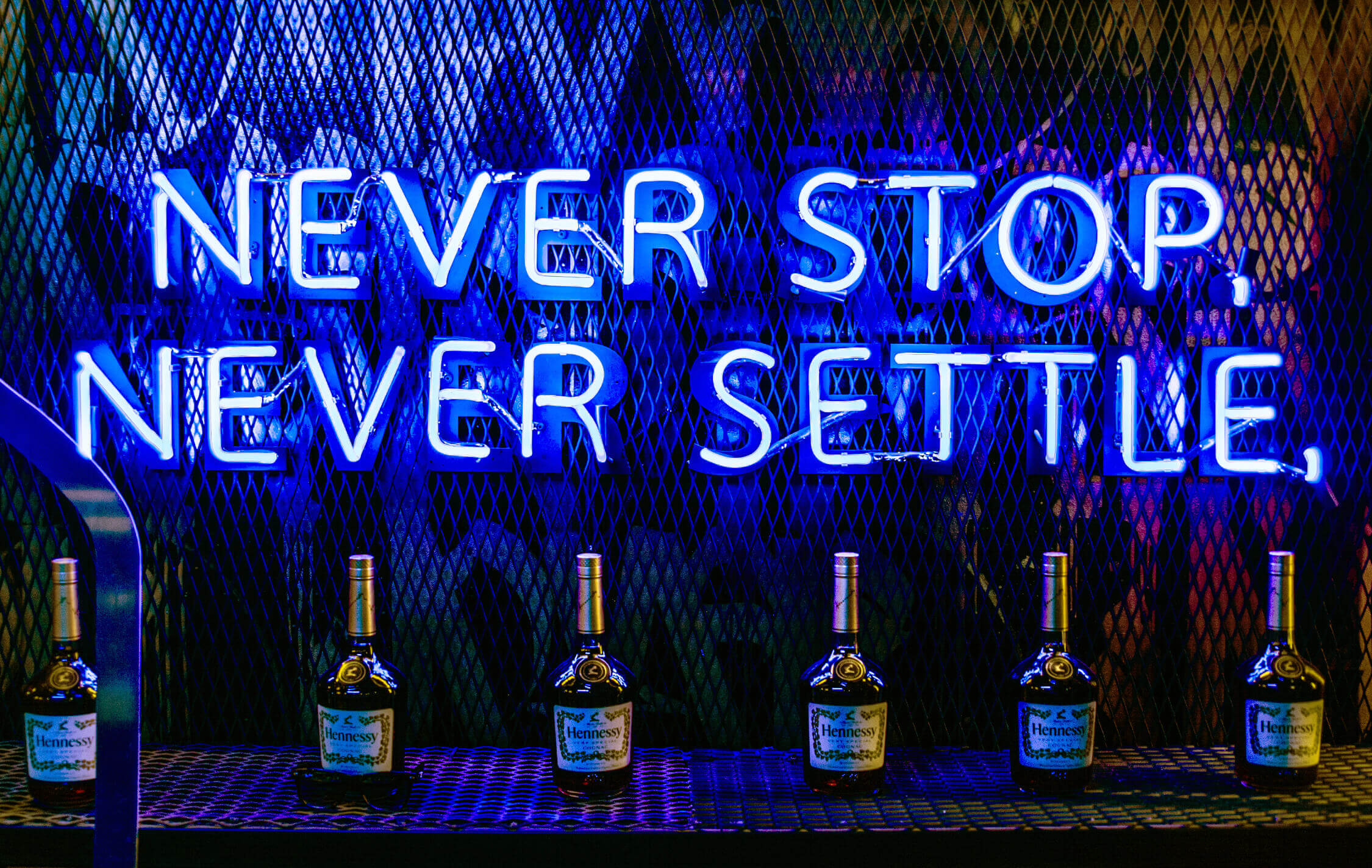 nie-aufhören-zu-siedeln-Hennessy-Neon-Werbung-Hennessy-Neon-Schriftzug-Neon-Aufschrift-auf-der-Brust-der-Flaschen-Blau-Neon-hinter-der-Bar