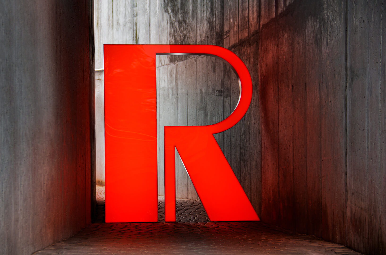 Lettre R grand format en rouge sur un mur en béton, éclairée par des LED.