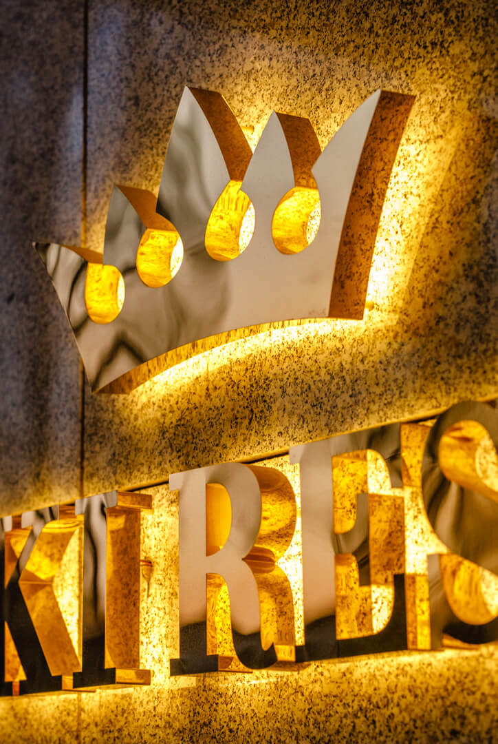 Krone im Logo, aus rostfreiem Stahl in Gold, hinterleuchtet auf LED-Wand.