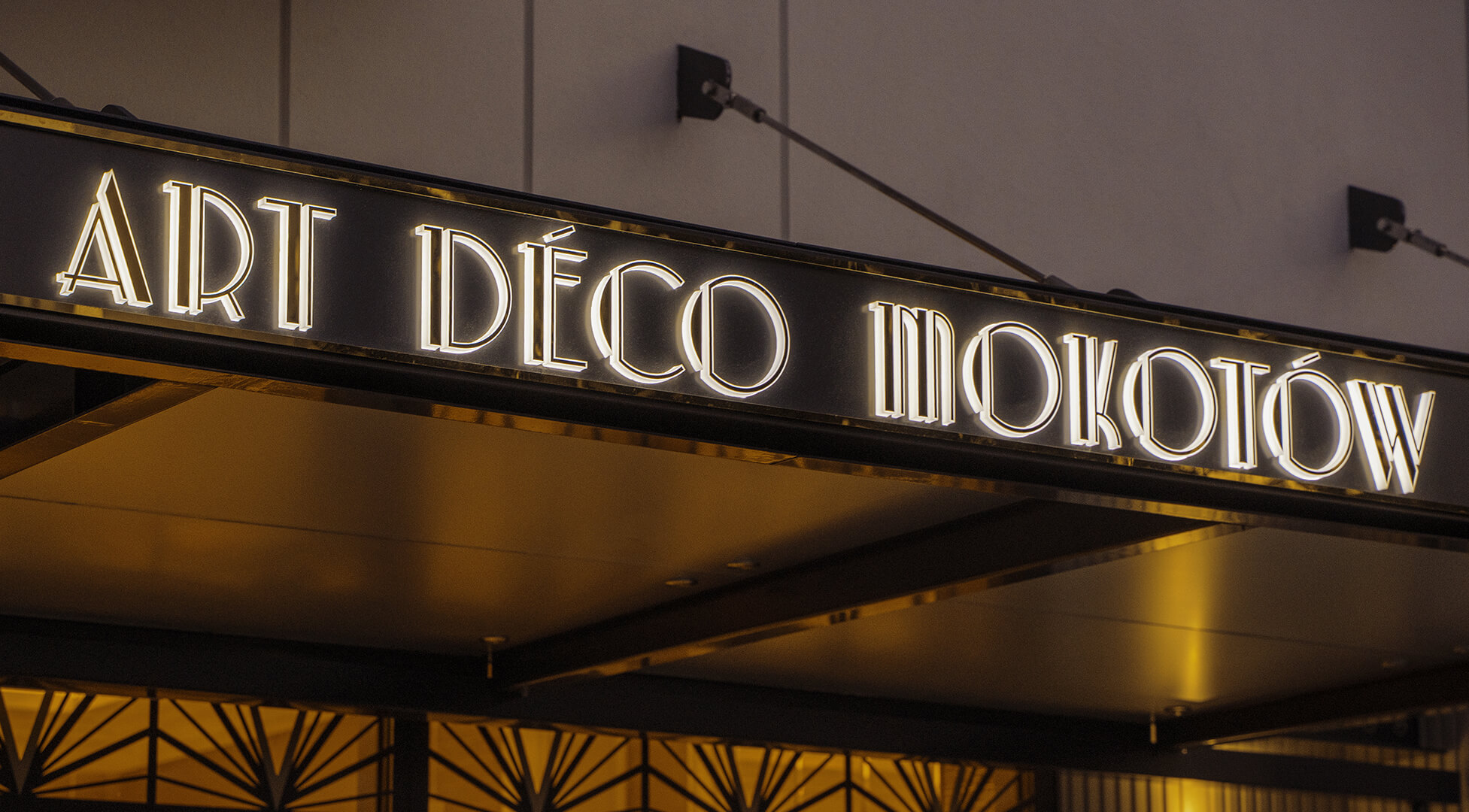 Art Deco Mokotow - Goldfarbene Dibond-Kassette über dem Mokotow-Eingang im Art-Déco-Stil, mit LED-Beleuchtung.