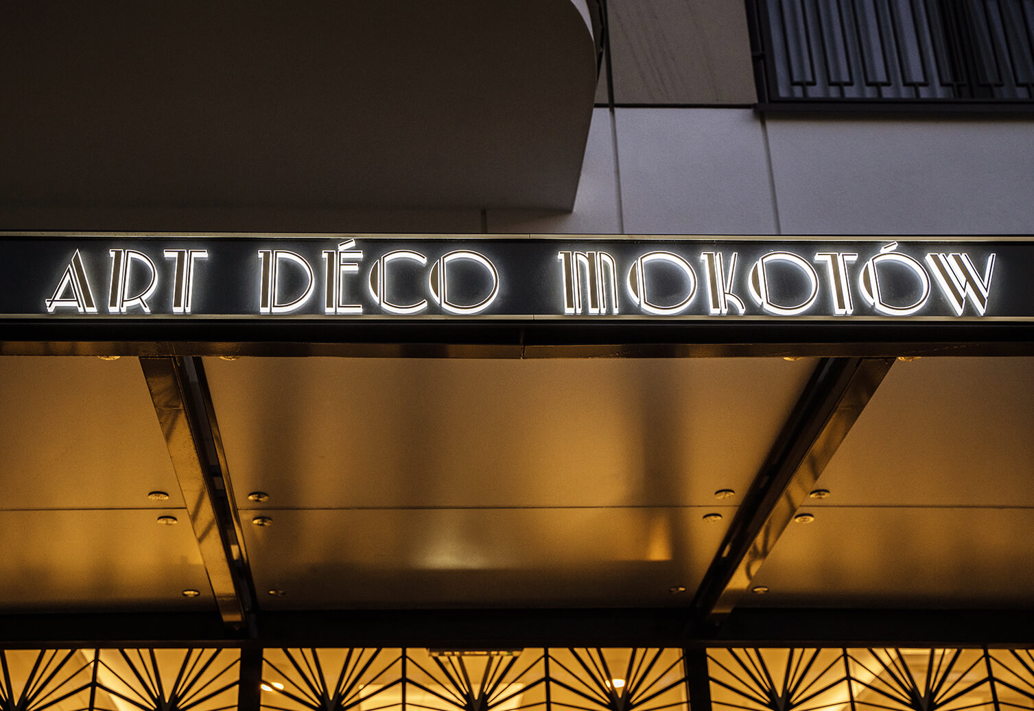 Art Deco Mokotów - Goldfarbene Dibond-Kassette über dem Mokotow-Eingang im Art-Déco-Stil, mit LED-Beleuchtung.
