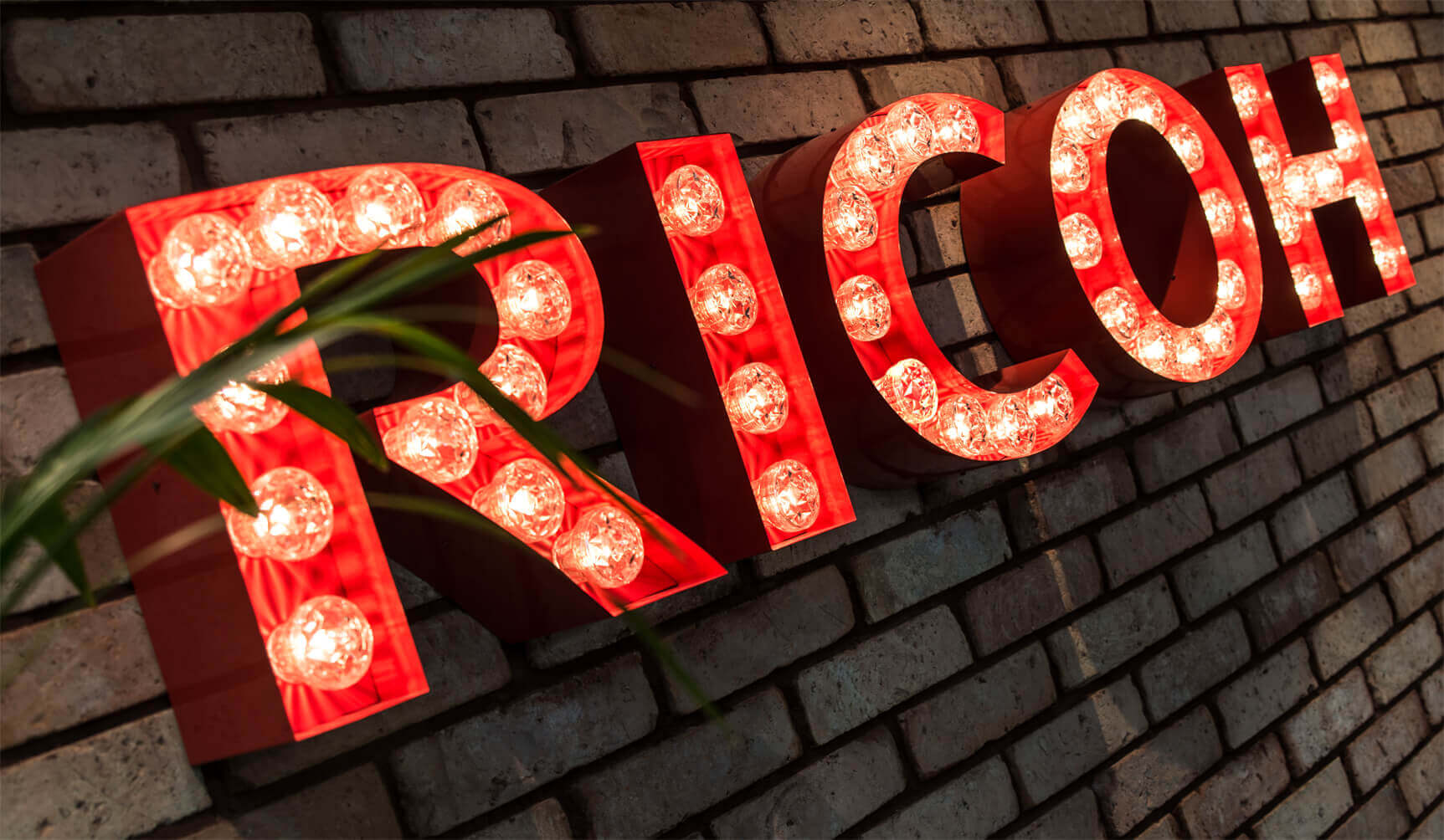 RICOH - RICOH - Buchstaben mit Glühbirnen an einer Backsteinmauer